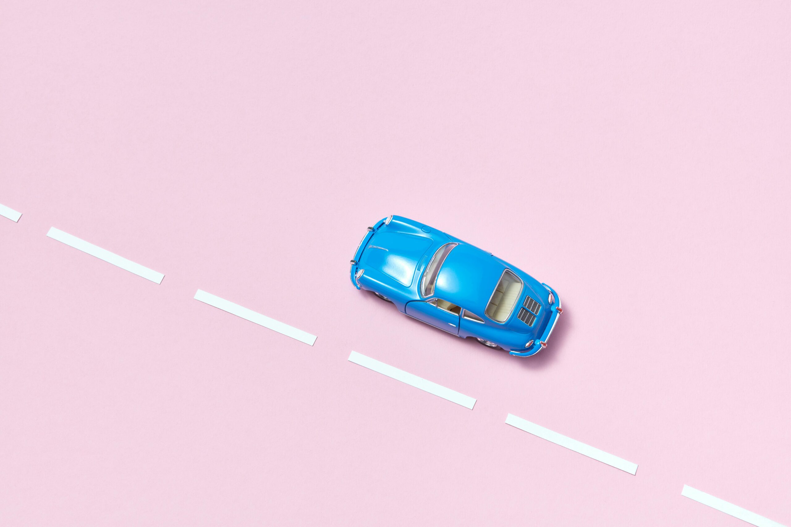 Minimalist image of a blue toy car on a pink road with white stripes, viewed from above.