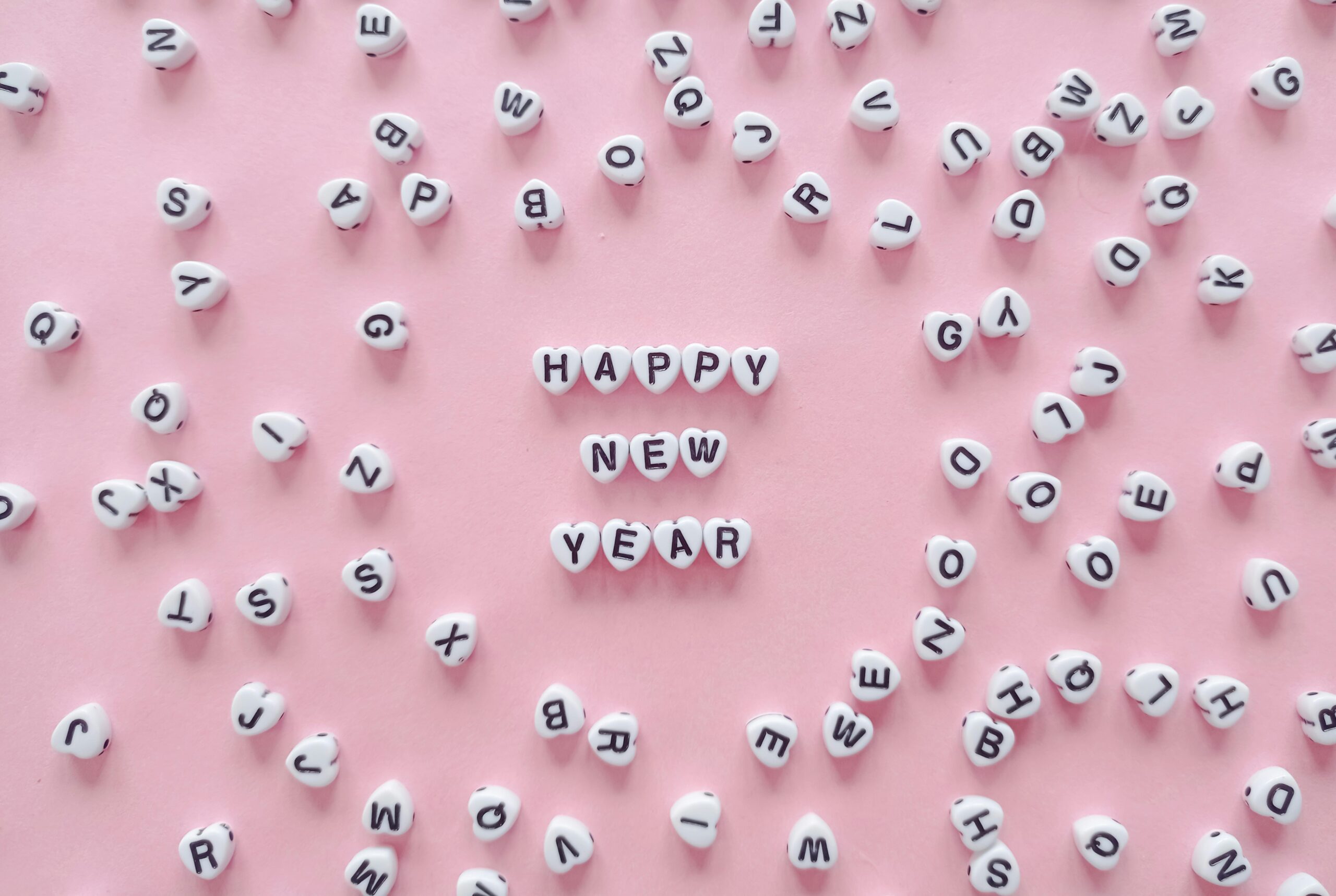 Heart-shaped letter tiles form 'Happy New Year' on a pink background. Minimalist and festive.