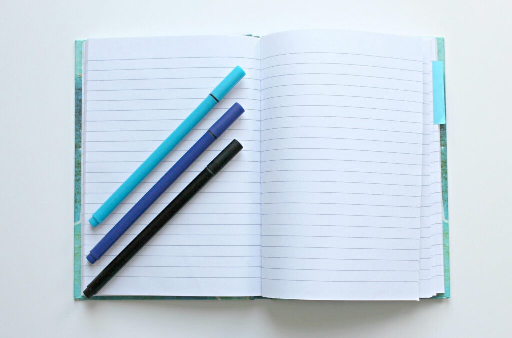 Open lined notebook with colorful pens on a white surface, perfect for note-taking and office tasks.