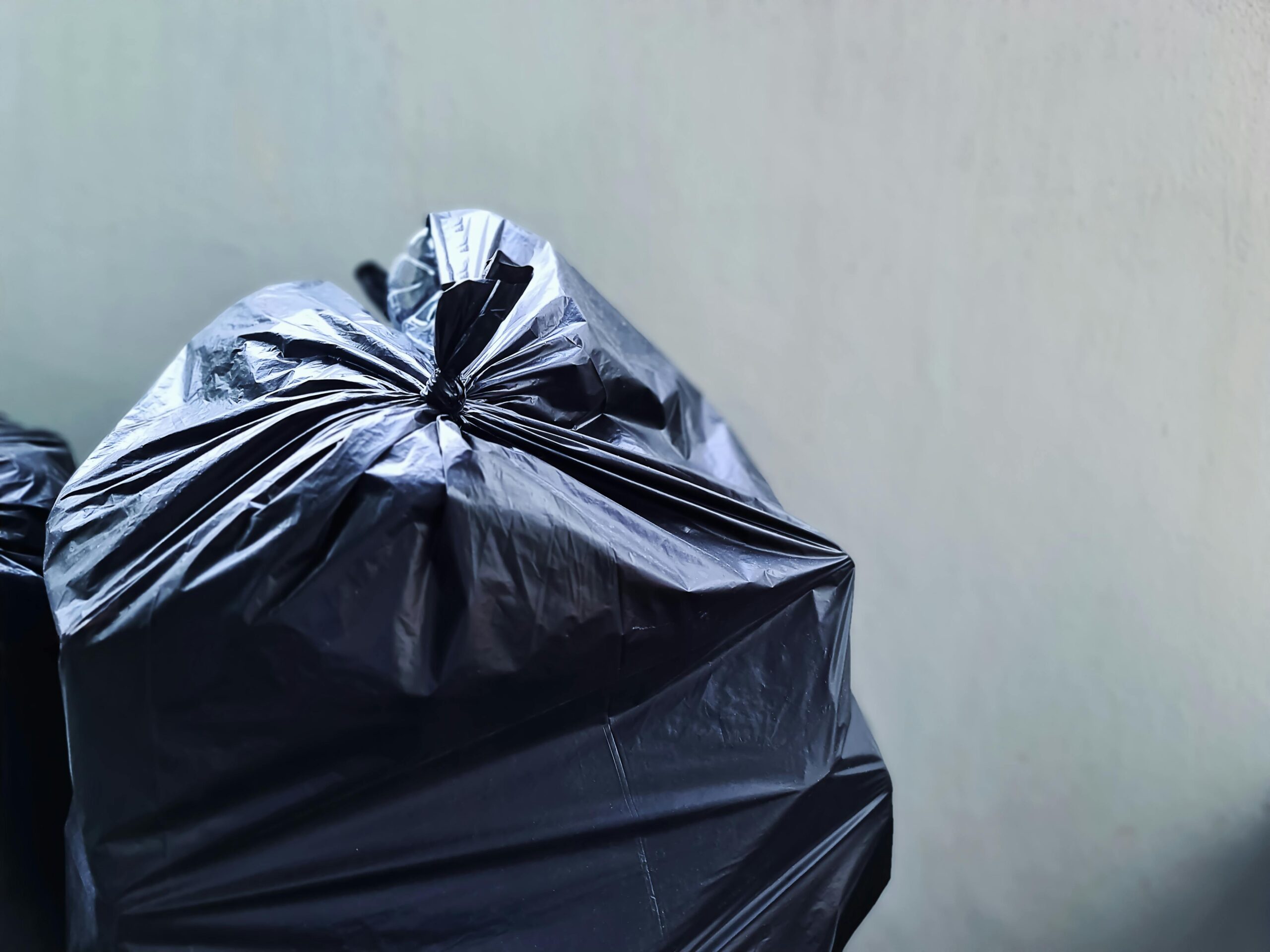 A close-up photograph of a black plastic garbage bag against a plain concrete wall, highlighting waste management.
